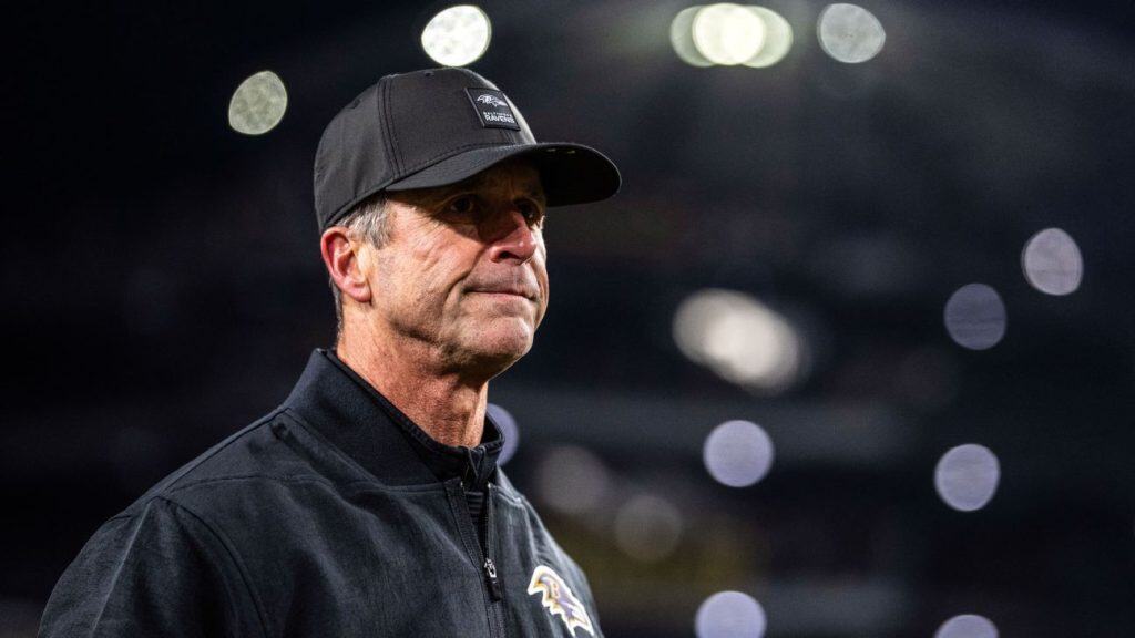 John Harbaugh, NFL coach in a black Baltimore Ravens jacket and cap looks ahead with a serious expression under bright stadium lights, with the football field and crowd softly blurred behind him.