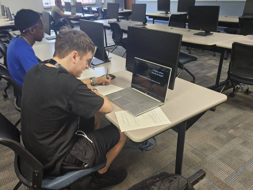 Students working on laptops in a modern classroom setting with multiple screens.