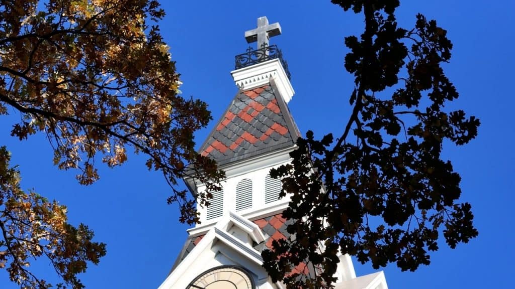 Church steeple with cross against a clear blue sky, surrounded by autumn leaves.