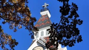 Church steeple with cross against a clear blue sky, surrounded by autumn leaves.