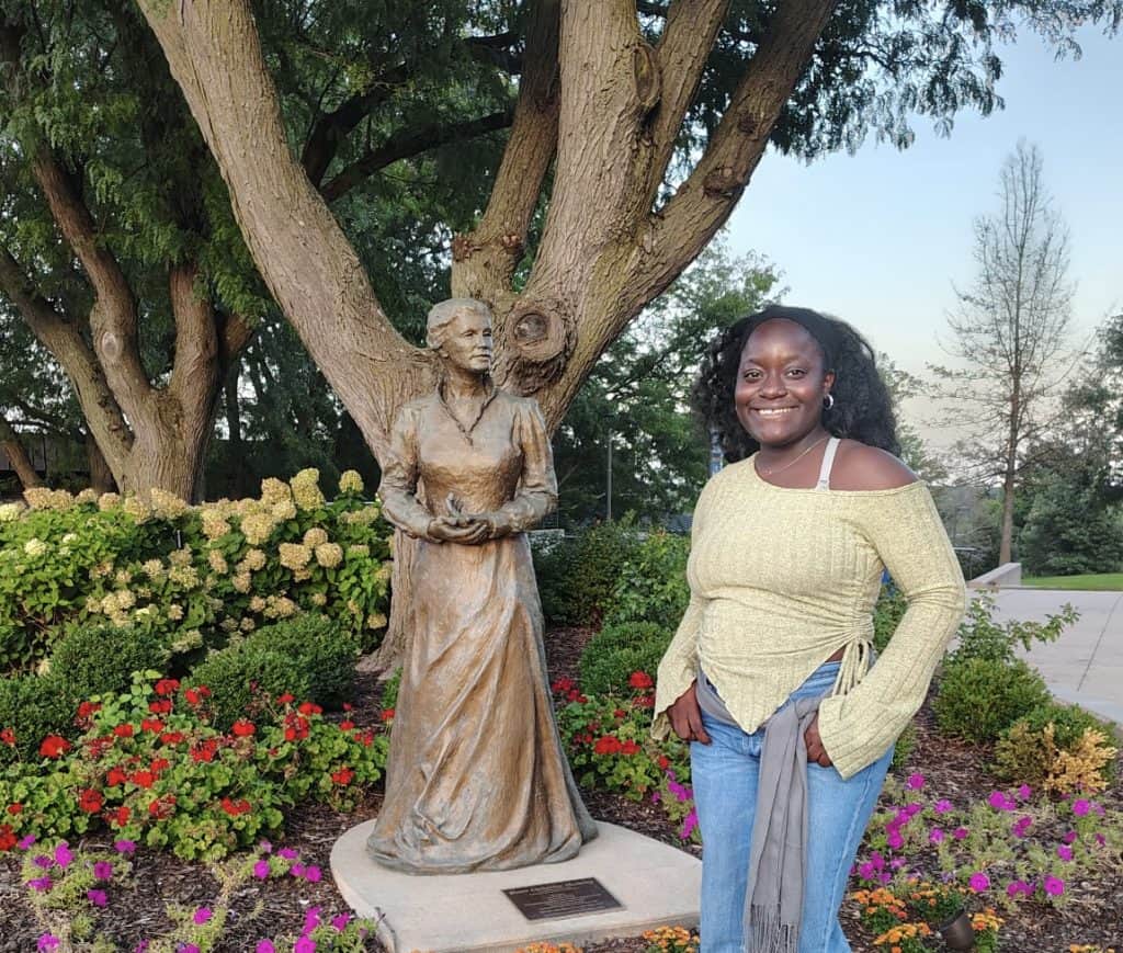 Young woman standing beside a statue of a woman in a garden with trees and flowers.