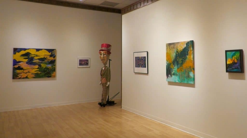 Art gallery with colorful paintings and a mannequin in a red hat.